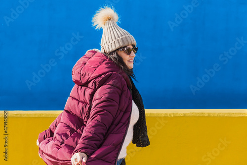 Portrait of Woman with smiling face with sunglasses purple jacket and woolen cap on blue and yellow background - Outdoor winter vacation concept.