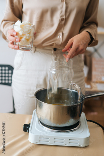 Fototapeta Naklejka Na Ścianę i Meble -  Vertical front view of female artisan adding white dry soy wax in glass jar into pot of boiling water for creating candle building mixture. Process of making handmade natural candle at workshop.