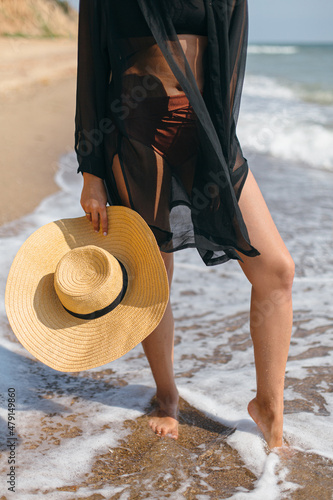 Stylish hat in hands of carefree woman walking in sea waves on sunny beach, close up. Summer vacation. Young fashionable female with straw hat relaxing on tropical shore. Carefree