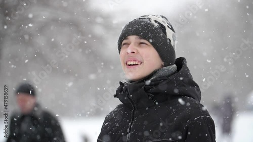 A 13-year-old European boy, dressed in a black jacket, stands outside in a heavy snowfall, he looks at the snow and smiles. Close-up, slow motion, shallow depth of field