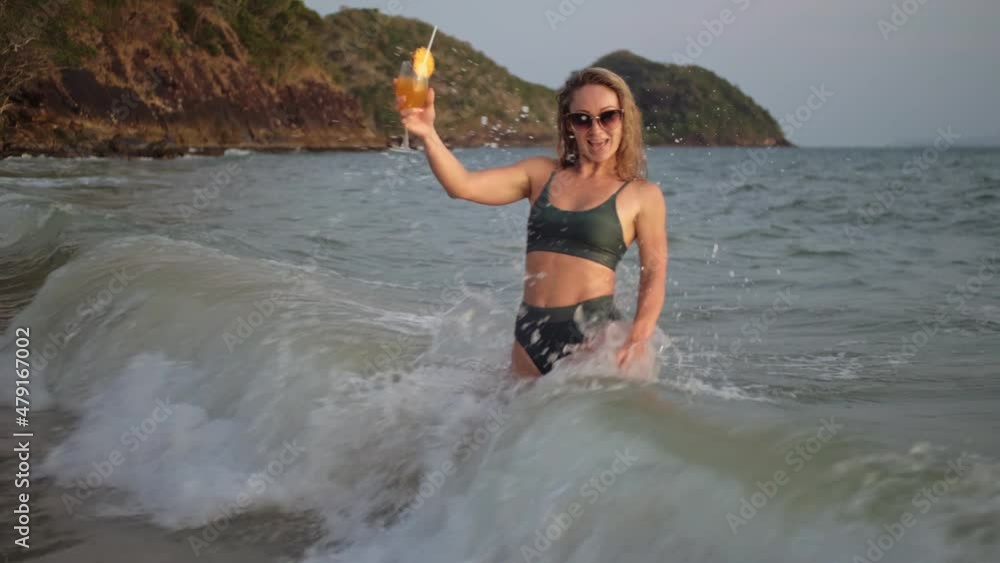 Woman stand knee-deep in sea on sunset. Girl on beach in green swimsuit having fun, drinks her orange cocktail, big waves hitting legs Woman stand knee-deep in sea on sunset. Girl on beach in green swimsuit having fun, drinks her orange cocktail, big waves hitting legs