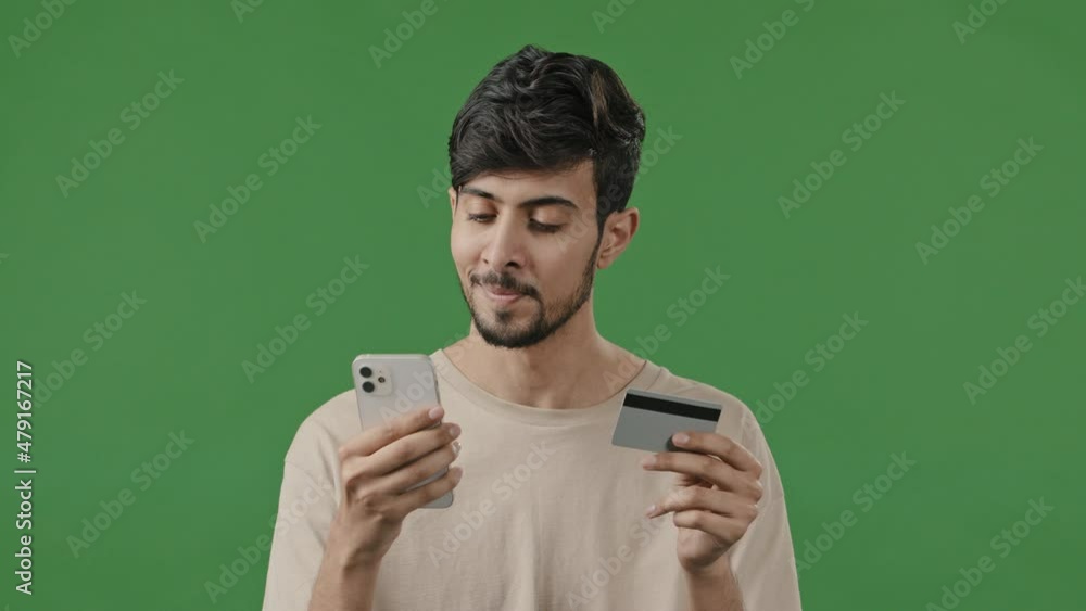 Happy smiling guy on green background in studio arab man pays internet order using phone and credit card enters number for transaction in app young male use mobile banking application remote shopping Happy smiling guy on green background in studio arab man pays internet order using phone and credit card enters number for transaction in app young male use mobile banking application remote shopping