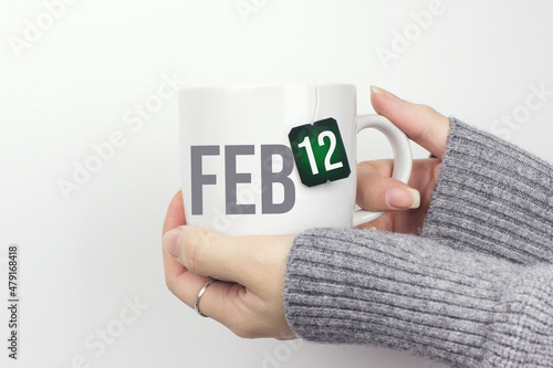 February 12nd. Day 12 of month, Calendar date. Closeup of female hands in grey sweater holding cup of tea with month and calendar date on teabag label. Winter month, day of the year concept. photo