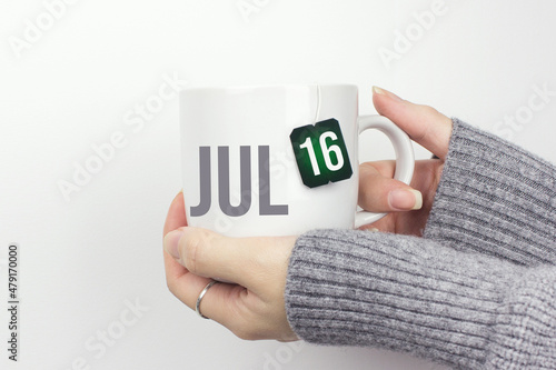 July 16th. Day 16 of month  Calendar date. Closeup of female hands in grey sweater holding cup of tea with month and calendar date on teabag label. Summer month  day of the year concept.