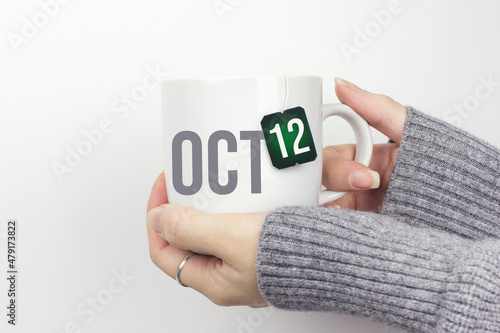 October 12nd. Day 12 of month, Calendar date. Closeup of female hands in grey sweater holding cup of tea with month and calendar date on teabag label. Autumn month, day of the year concept. photo