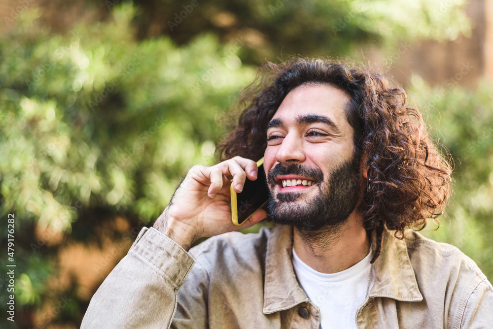 Glad bearded Hispanic man with dark curly hair looking away with smile. He is answering phone call on blurred background of lush tree branches in daytime Glad bearded Hispanic man with dark curly hair looking away with smile. He is answering phone call on blurred background of lush tree branches in daytime