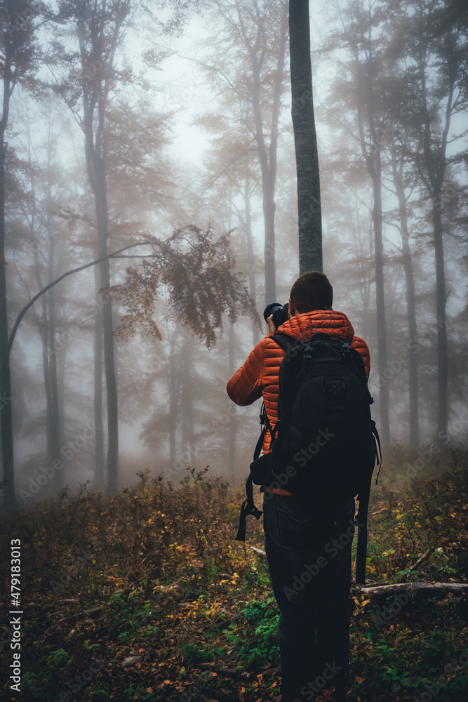 Young bearded hiker wearing orange jacket taking photos at forest with fog and mist. Back view of photographer in field enjoying the view.