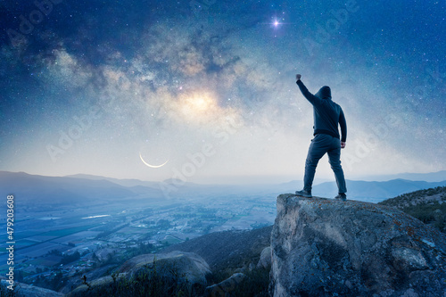 man standing on the top of the mountain, back view, with a hand up celebrating and Milky Way over the valley.