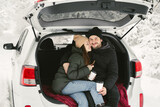 Young couple, a man and a woman, are sitting in the trunk of a car in a winter, snowy forest, hugging, kissing and drinking coffee