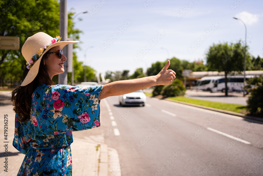 Profile of a woman in floral summer dress and hat hitchhiking next to a street