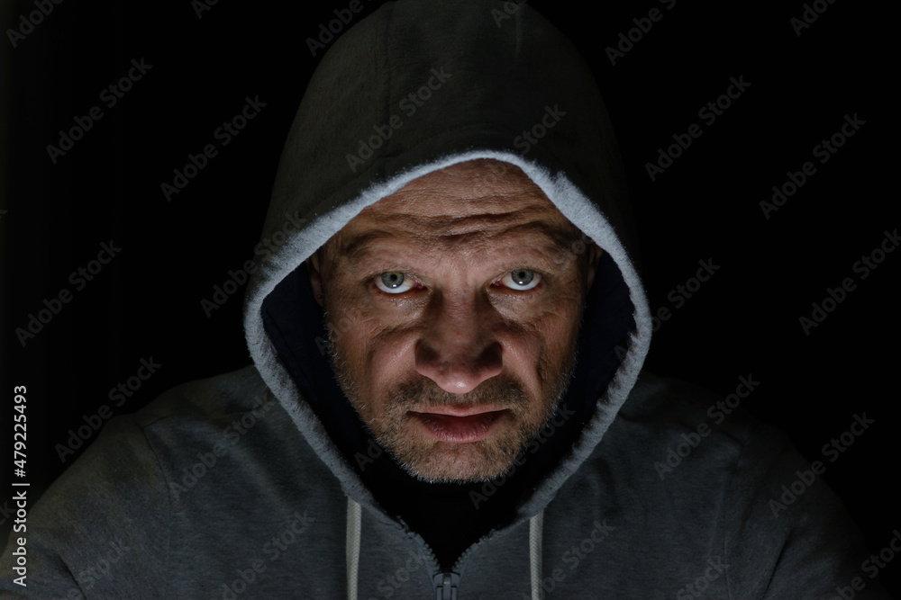 A man in a hood, a lighted face on a dark background. Evil grimace, low key. A man in a hood, a lighted face on a dark background. Evil grimace, low key.
