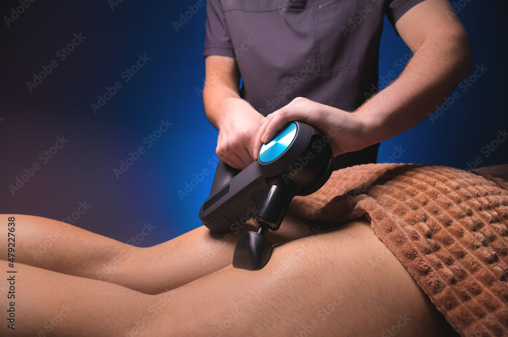 Male masseur doing massage exercises on the thighs with the help of a percussion vibro massager to a male client. Shock therapy for a regenerating massage of the athletic body.