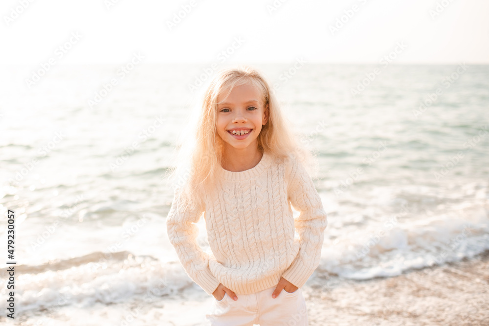 Happy smiling child girl 7-8 year old wear white knitted sweater and pants posing over nature sea backgorund. Funny blonde kid laughing walk at beach outdoor. Looking at camera. Summer vacation season