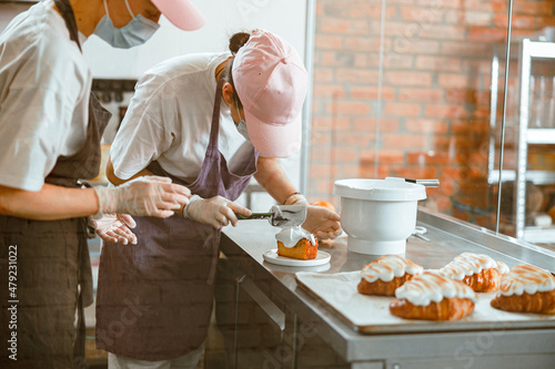 Принти на полотні Confectioner trainee spreads cream onto sponge cake while colleague teaches her