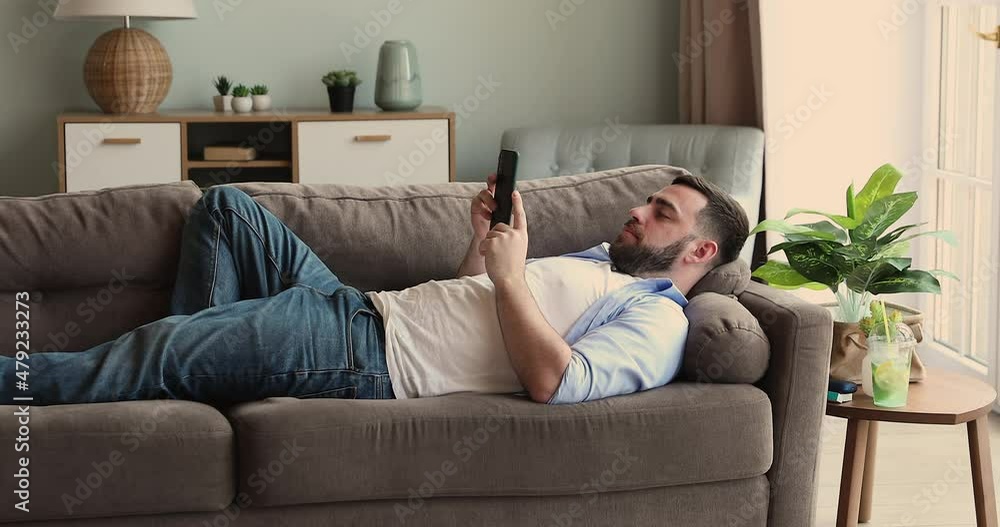 Relaxed millennial guy lie on back on soft comfortable couch at living room spend lazy weekend watching video on mobile phone screen. Calm young man rest use cellphone to check email read news online Relaxed millennial guy lie on back on soft comfortable couch at living room spend lazy weekend watching video on mobile phone screen. Calm young man rest use cellphone to check email read news online