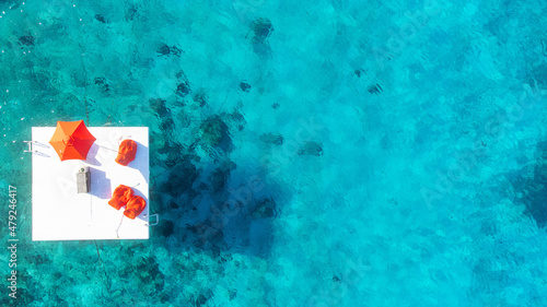 A white dock with vibrant red umbrella and furniture is surrounded by turquoise blue Caribbean sea water. The ocean is clear and coral reefs can be seen below. Aerial vew by drone. 