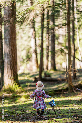 Toddler girl from behind wearing a hat and Easter dress holding an Easter basket hunting for Easter eggs in a forest.