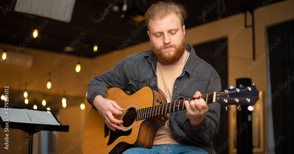 Obraz premium A young guy with a beard plays an acoustic guitar in a room with warm lighting