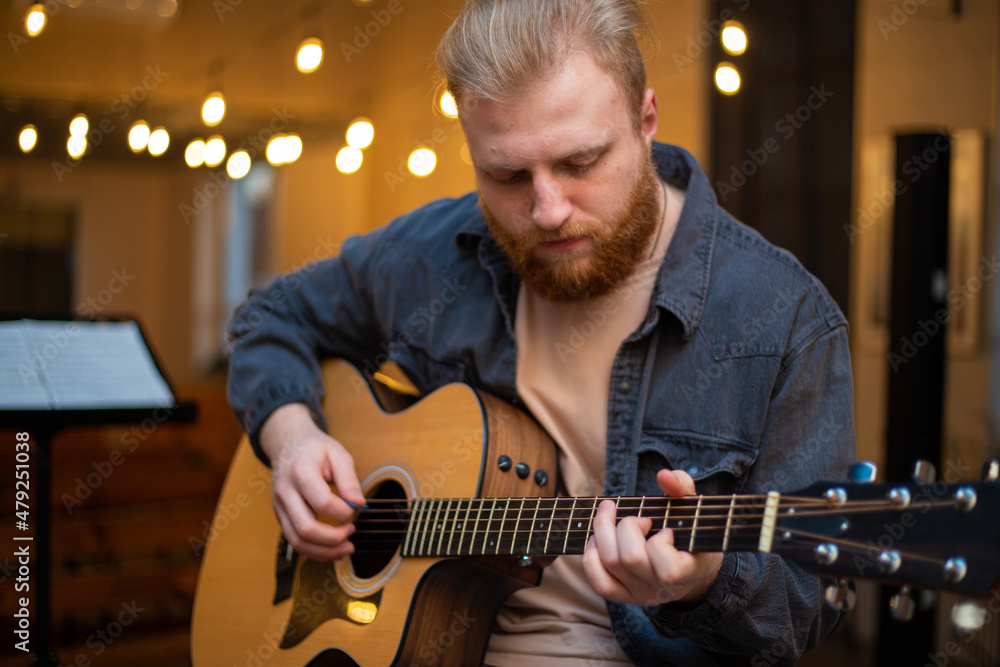 Obraz premium A young guy with a beard plays an acoustic guitar in a room with warm lighting