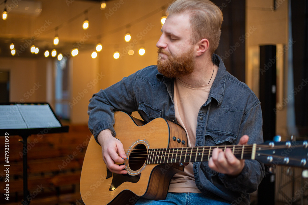 Obraz premium A young guy with a beard plays an acoustic guitar in a room with warm lighting