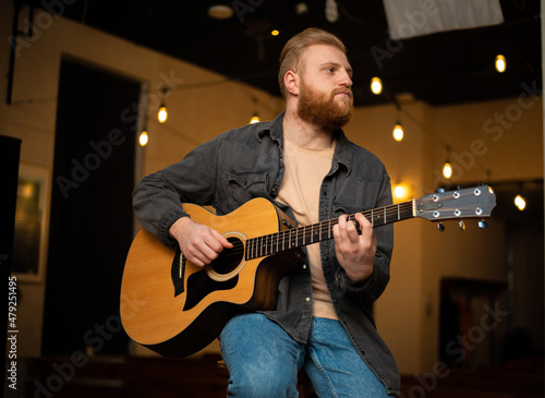 A young guy with a beard plays an acoustic guitar in a room with warm lighting