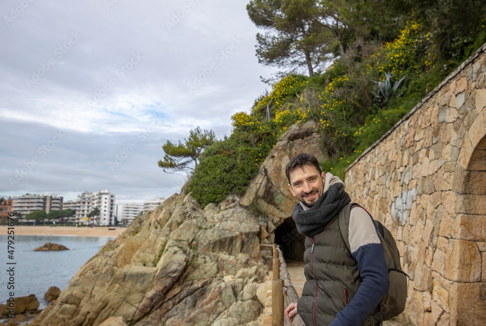 Handsome man smiling on the beach on an autumn day. A man in warm clothes stands near a stone cliff.