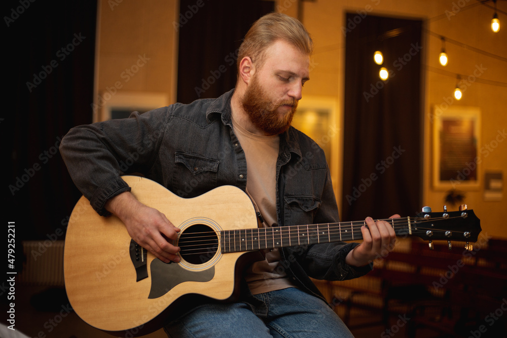 Obraz premium A young guy with a beard plays an acoustic guitar in a room with warm lighting
