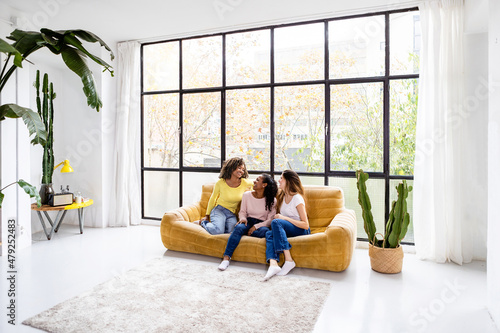 Indoor shot of three happy young women sitting on sofa - Young multiracial female friends hanging out and relaxing at home - Social gathering, friendship, youth and millennial people concept