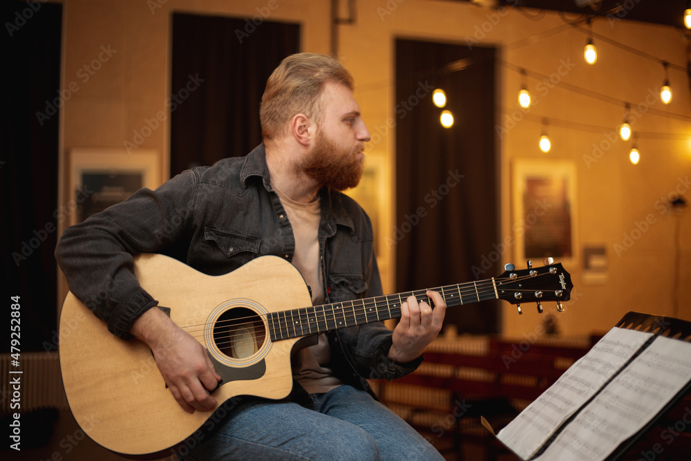 Obraz premium A young guy with a beard plays an acoustic guitar in a room with warm lighting