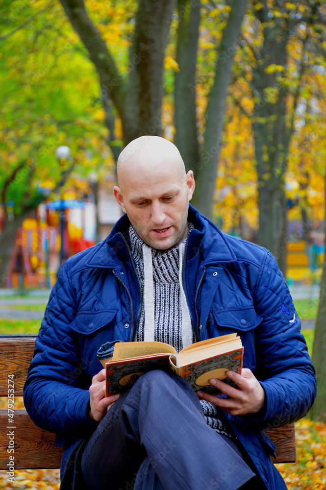 Fototapeta premium young man reads a book outdoors, sitting on a bench in an autumn park. serenity, relaxation and privacy concept