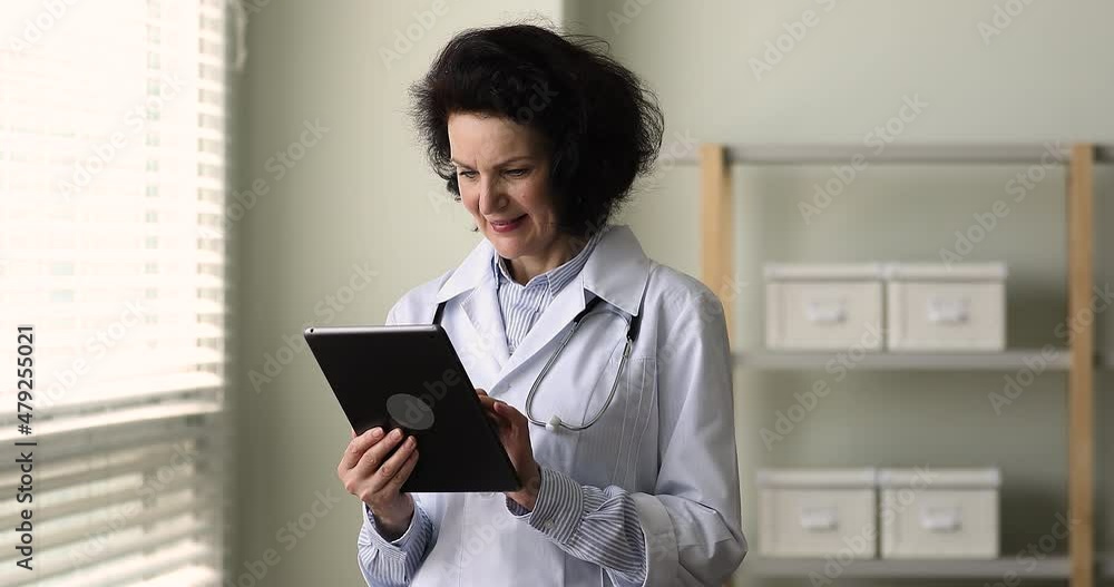 Happy elderly female doctor using health-care app on digital tablet, standing in clinic office smile enjoy easy comfort modern tech usage, web surfing medical info, gives on-line consultation concept Happy elderly female doctor using health-care app on digital tablet, standing in clinic office smile enjoy easy comfort modern tech usage, web surfing medical info, gives on-line consultation concept