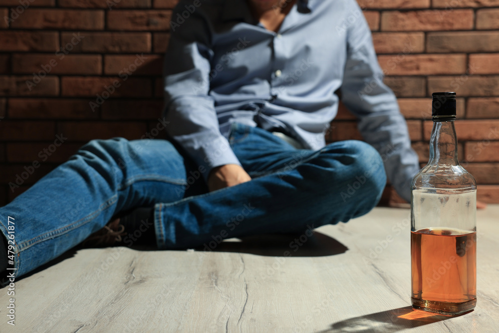 Addicted man sitting near red brick wall, focus on bottle of alcoholic drink Addicted man sitting near red brick wall, focus on bottle of alcoholic drink