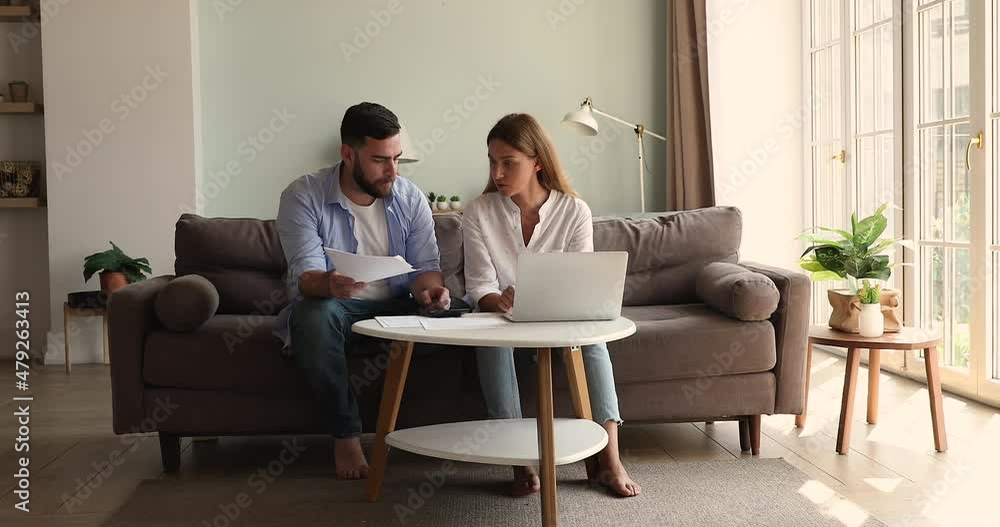 Focused young spouses work with papers at cozy home office manage family business sitting on sofa at living room. Concentrated husband wife review insurance payment check bills before pay online on pc Focused young spouses work with papers at cozy home office manage family business sitting on sofa at living room. Concentrated husband wife review insurance payment check bills before pay online on pc