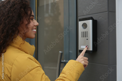 Fototapeta Young African-American woman ringing intercom with camera near building entrance