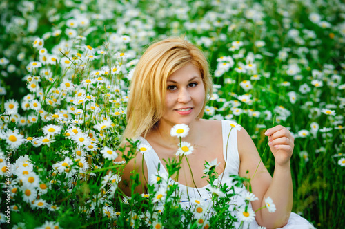 Portrait of a young, plump, beautiful woman resting on a chamomile field at sunset. A plus-size woman in a white sundress sits in a meadow with daisies.