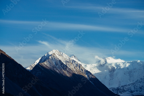 Atmospheric alpine landscape with sunlit snow-covered mountain top under cirrus clouds in blue sky. Awesome scenery with beautiful pointy peak with snow and high snowy mountain wall with low clouds.