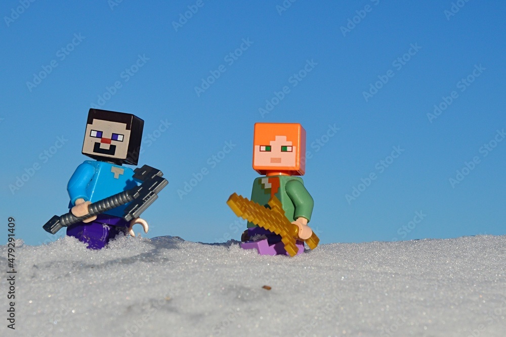 Naklejka premium LEGO Minecraft figures of Steve with iron pickaxe and Alex with golden sword standing knee deep in real snow sunny winter day, blue skies in background. 