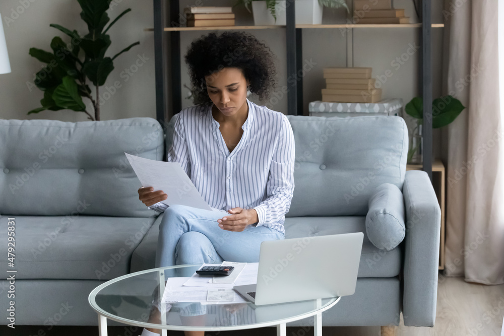 Focused millennial generation african ethnicity multiracial woman holding paper bill in hands, checking domestic utility payments, planning investments or making financial audit using computer at home Focused millennial generation african ethnicity multiracial woman holding paper bill in hands, checking domestic utility payments, planning investments or making financial audit using computer at home