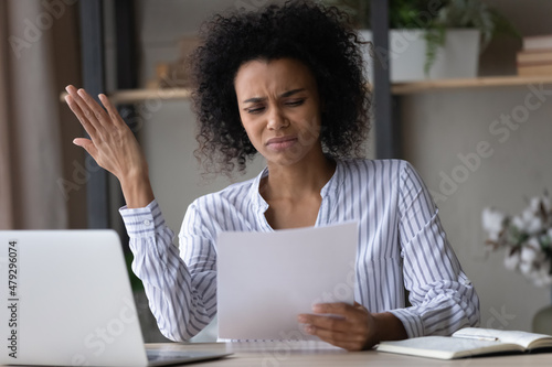 Valokuva Stressed young african american mixed race businesswoman looking at paper document in hands, feeling dissatisfied with bad research results, finding mistakes in financial report, confused with letter