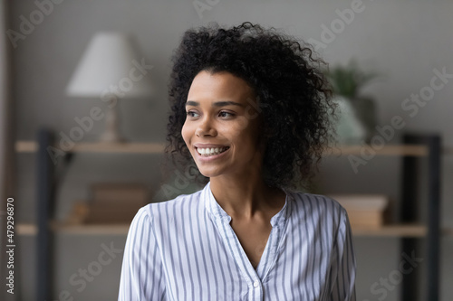 Happy dreamy beautiful curly motivated young african american multiracial woman looking in distance, daydreaming alone or visualizing future standing in modern living room, inspiration concept.