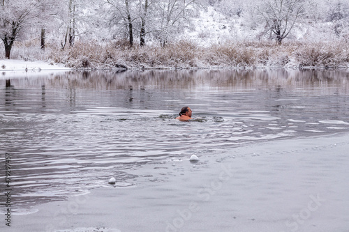 A middle-aged man swims on a cold winter day along the river against the backdrop of snow-covered trees. The concept of hardening, healthy lifestyle