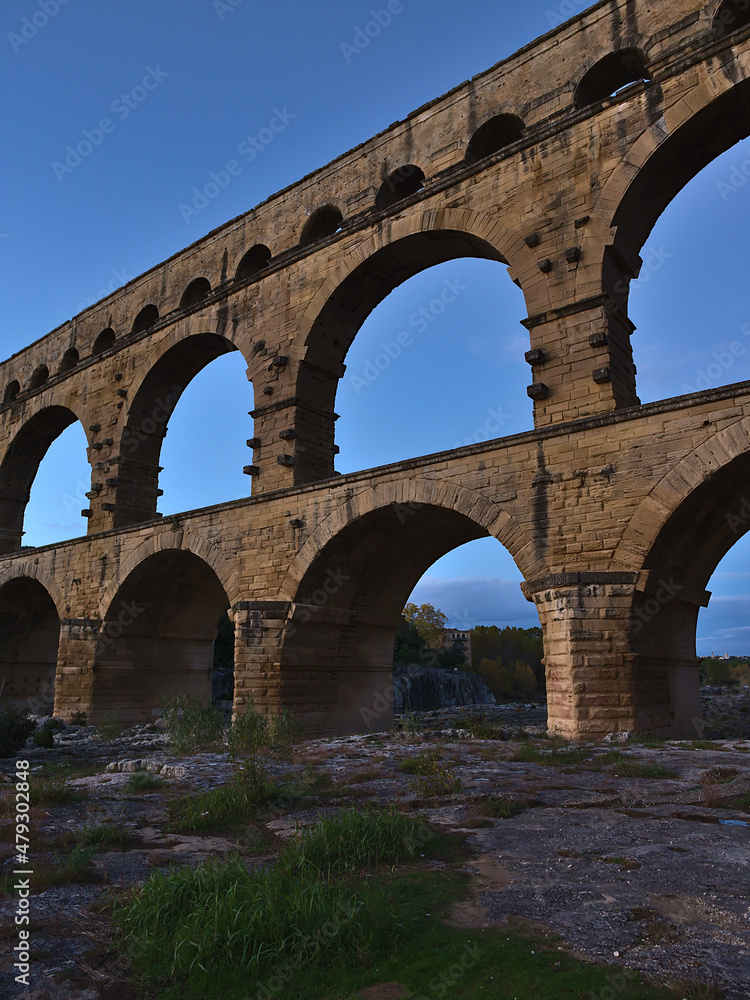 View of ancient Roman aqueduct Pont du Gard with majestic stone pillars above Gardon river after sunset near Vers-Pont-du-Gard, Occitanie, France.