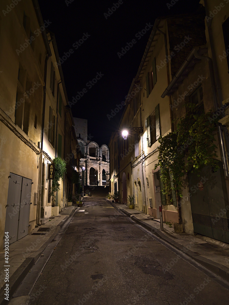 Night view of narrow alley in historic center of town Arles, Provence, France with old traditional buildings, street lights and Roman amphitheatre.