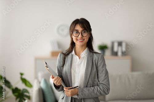 Portrait of cheerful female psychologist posing with clipboard, looking and smiling at camera, working in modern office photo