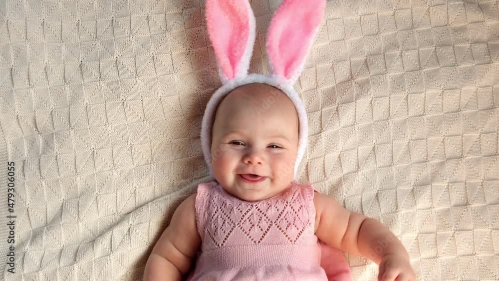Funny little Child is wearing Bunny Ears on Cozy Background. Happy Easter. Baby Girl in pink Dress and Headband with Ears is lying on a Bed. Cute Newborn Baby Smiling and Looking to the Camera. Funny little Child is wearing Bunny Ears on Cozy Background. Happy Easter. Baby Girl in pink Dress and Headband with Ears is lying on a Bed. Cute Newborn Baby Smiling and Looking to the Camera.