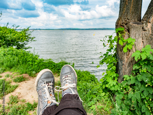 The girl sits by the lake, wears on her feet sneakers. Female legs