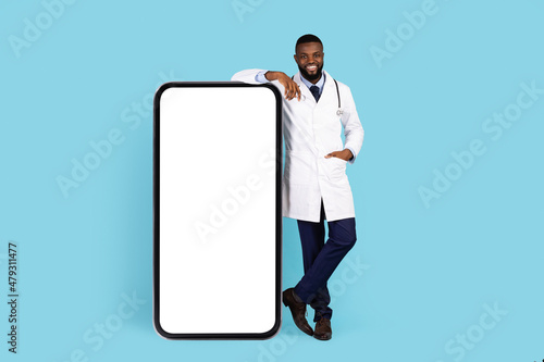 Medical Advertisement. Handsome Black Doctor In Uniform Standing Near Big Blank Smartphone
