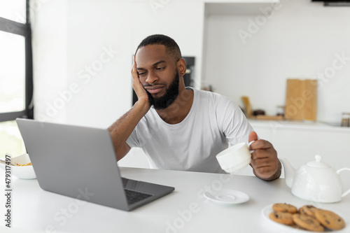 Fotografie Tired black man using laptop sitting at kitchen table
