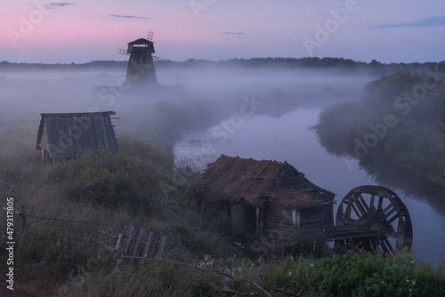 Sunset in the old village. A dilapidated water mill with a thatched roof is located on the banks of the river. In the background, a village windmill hidden in the fog. Summer landscape with a river.  photo