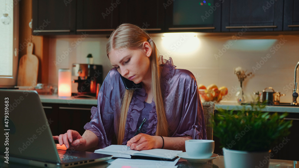 Young business woman working at home by sitting at the computer and speaking by smartphone. She is in home bathrobe in the kitchen.